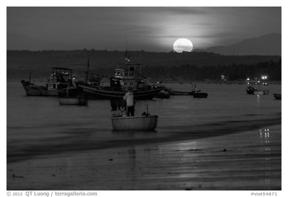 Fisherman paddling on coracle boat towards fishing boats at moonset. Mui Ne, Vietnam (black and white)