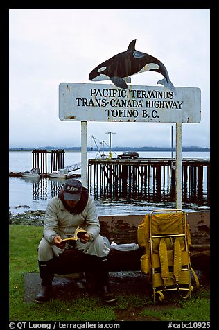 Backpacker sitting under the Transcanadian terminus sign, Tofino. Vancouver Island, British Columbia, Canada (color)