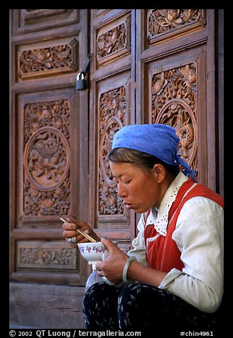 Bai woman eating from a bowl in front of carved wooden doors. Dali, Yunnan, China