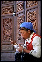 Bai woman eating from a bowl in front of carved wooden doors. Dali, Yunnan, China