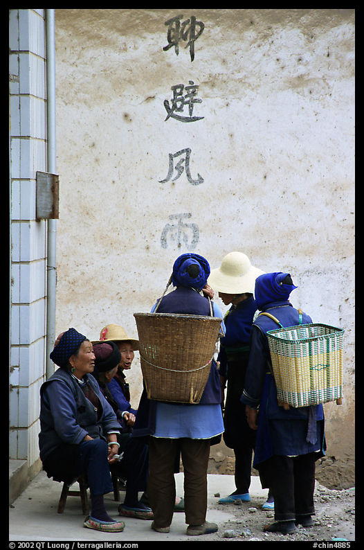 Elderly women with back baskets in front of a wall with Chinese scripture. Shaping, Yunnan, China