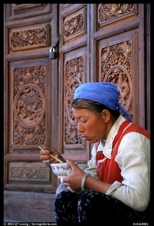 Bai woman eating from a bowl in front of carved wooden doors. Dali, Yunnan, China