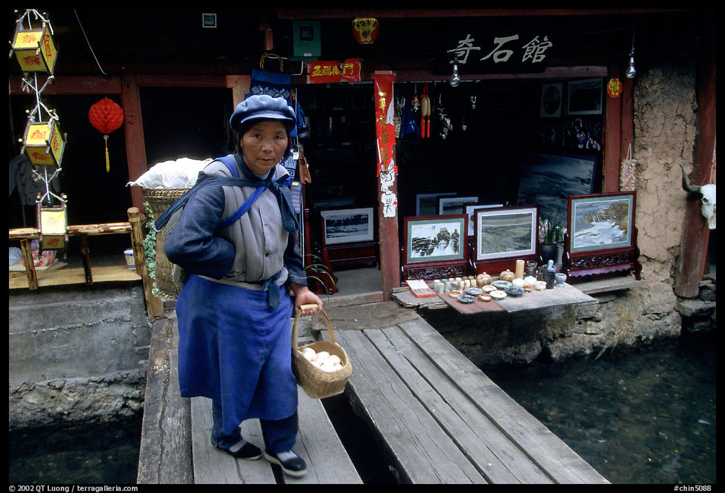 Naxi woman peddling eggs  to local residents walks acros a canal. Lijiang, Yunnan, China