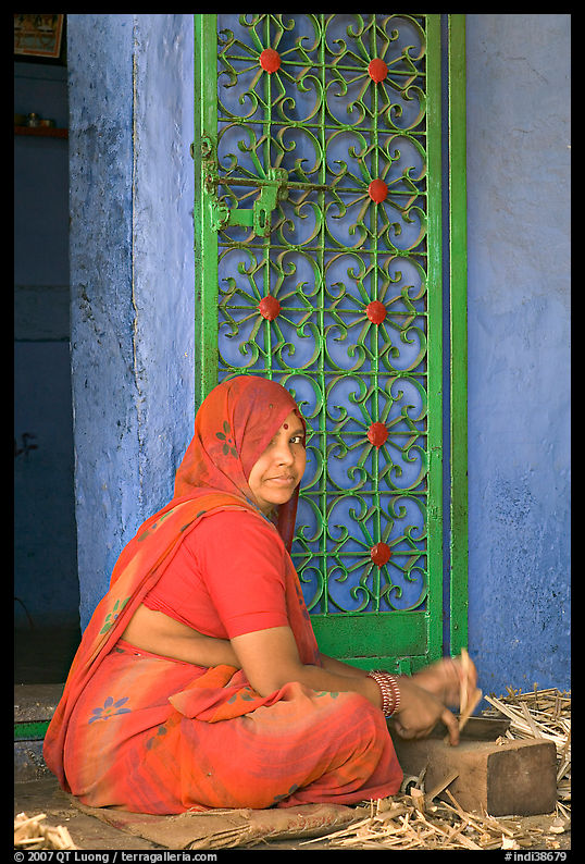 Woman in orange sari sitting next to green door and blue wall. Jodhpur, Rajasthan, India