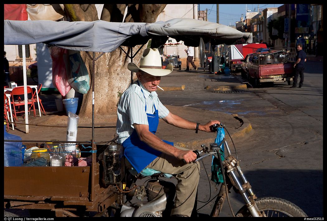 Man with cigarette riding a motorcycle-powered food stand on town plaza. Mexico