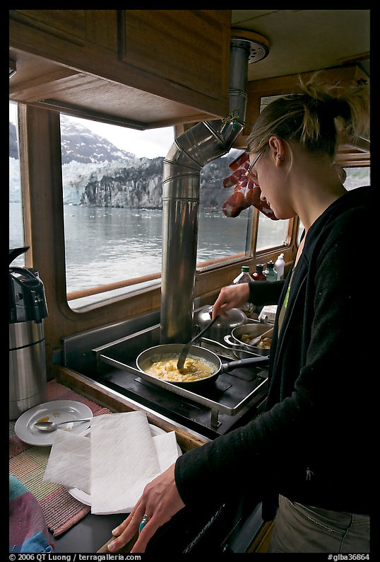 Woman cooking eggs aboard small tour boat, with glacier outside. Glacier Bay National Park, Alaska, USA.