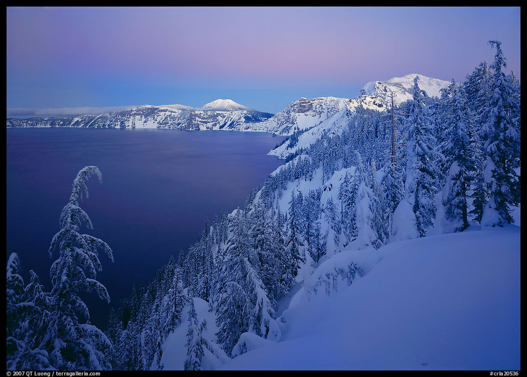 Snow-covered rim and trees, lake and mountains, dusk. Crater Lake National Park, Oregon, USA.
