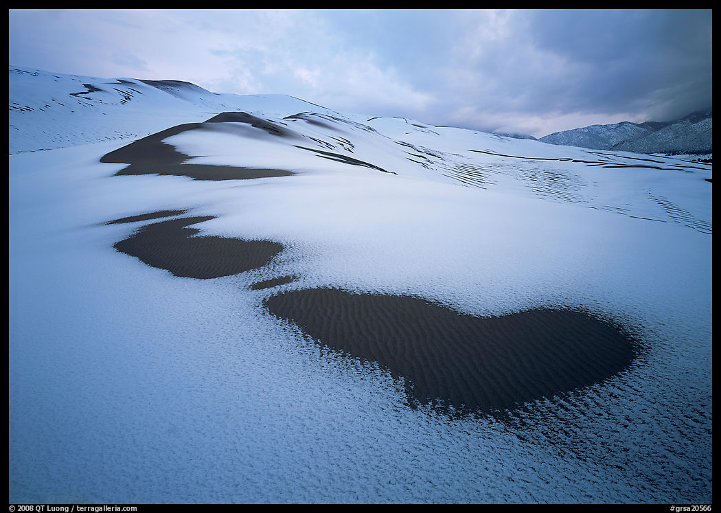 Patches of uncovered sand in snow-covered dunes, mountains, and dark clouds. Great Sand Dunes National Park, Colorado, USA.