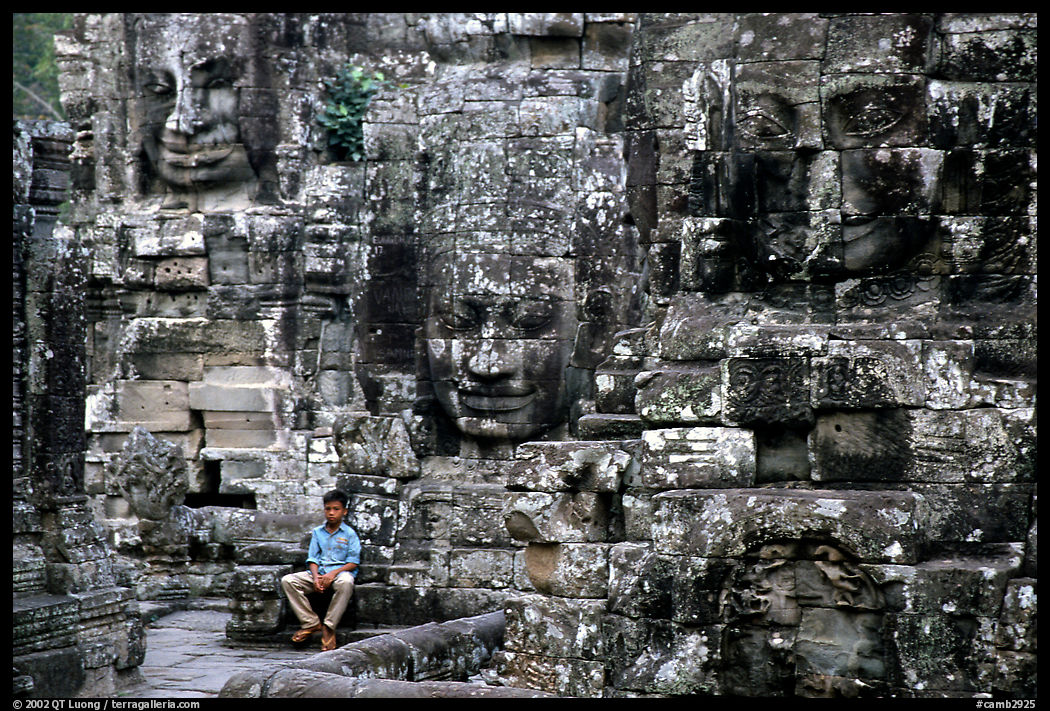 Boy sits next to large stone smiling faces, the Bayon. Angkor, Cambodia