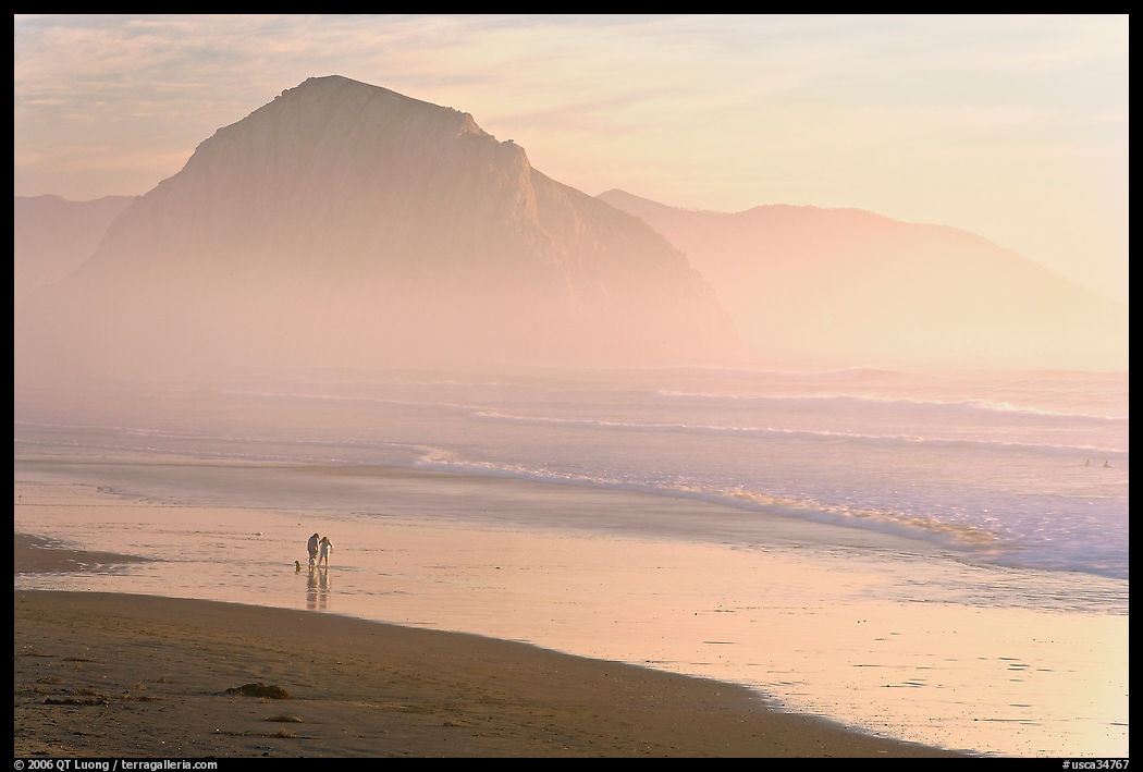 Couple and dog reflected in wet sand, with Morro Rock behind, sunset. Morro Bay, USA