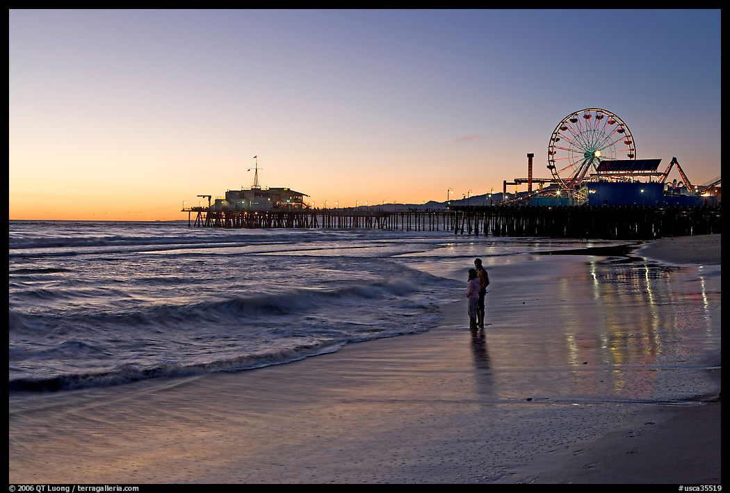 Couple reflected in wet sand at sunset, with pier and Ferris Wheel behind. Santa Monica, Los Angeles, California, USA