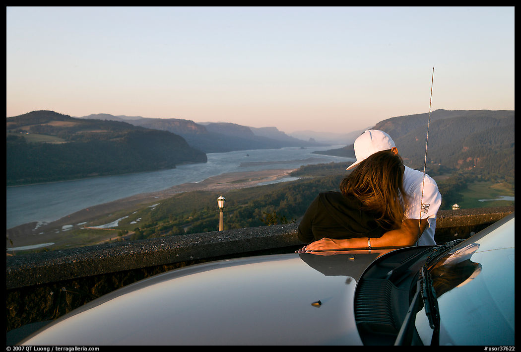 Couple embracing on car hood, with view of mouth of river gorge. Columbia River Gorge, Oregon, USA