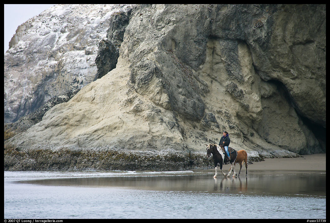 Woman horse-riding on beach next to sea cave entrance. Bandon, Oregon, USA