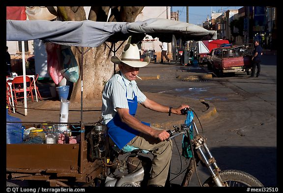 Man with cigarette riding a motorcycle-powered food stand on town plaza. Mexico