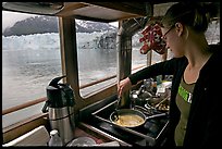 Woman cooking eggs aboard small tour boat, with glacier in view. Glacier Bay National Park, Alaska, USA.
