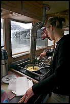 Woman cooking eggs aboard small tour boat, with glacier outside. Glacier Bay National Park, Alaska, USA. (color)