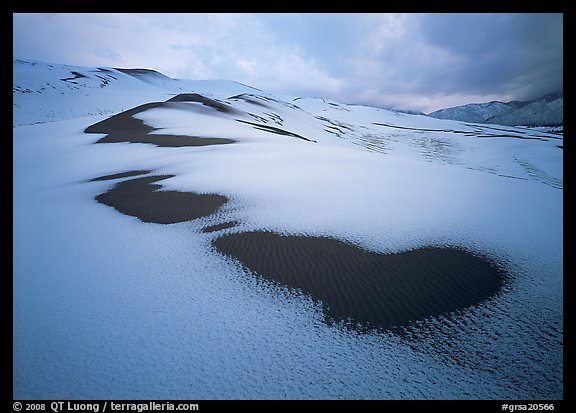 Patches of uncovered sand in snow-covered dunes, mountains, and dark clouds. Great Sand Dunes National Park and Preserve, Colorado, USA.