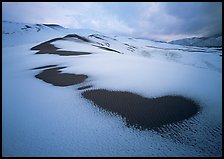 Patches of uncovered sand in snow-covered dunes, mountains, and dark clouds. Great Sand Dunes National Park and Preserve, Colorado, USA.