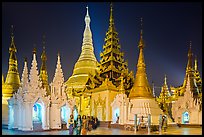 Women walking, stupas, shrines, and Main Stupa at night, Shwedagon Pagoda. Yangon, Myanmar