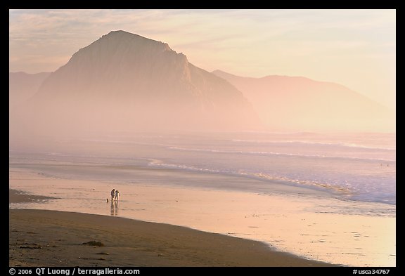 Couple and dog reflected in wet sand, with Morro Rock behind, sunset. Morro Bay, USA (color)