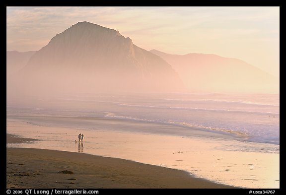 Couple and dog reflected in wet sand, with Morro Rock behind, sunset. Morro Bay, USA