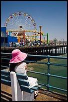 Woman sitting on bench with pink hat and ferris wheel. Santa Monica, Los Angeles, California, USA