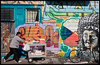 Man pushes vending cart pass mural and bicycle, Mission District. San Francisco, California, USA