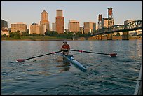 Woman rowing on racing shell and city skyline at sunrise. Portland, Oregon, USA