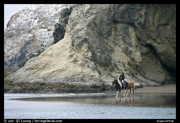 Woman horse-riding on beach next to sea cave entrance. Bandon, Oregon, USA (color)