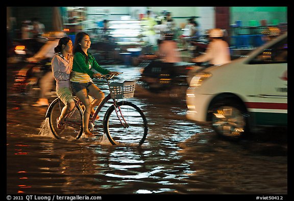Women sharing a bicycle ride at night on a water-filled street. Ho Chi Minh City, Vietnam