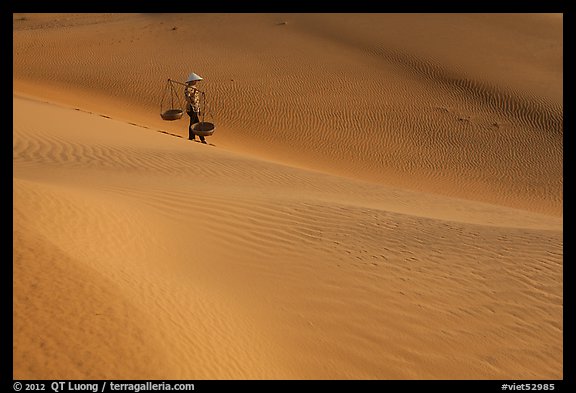 Woman with conical hat and yoke baskets pauses on sand dunes. Mui Ne, Vietnam (color)