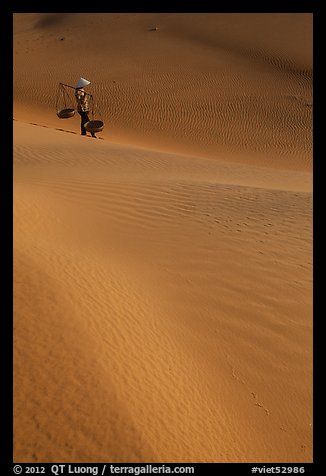 Woman with conical hat and shoulder pole baskets pauses on dune. Mui Ne, Vietnam (color)