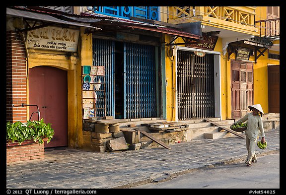 Woman carrying fruit in front of old storefronts. Hoi An, Vietnam (color)