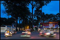 Blurred motorbikes at dusk and tall trees next to Van Hoa Park. Ho Chi Minh City, Vietnam (color)